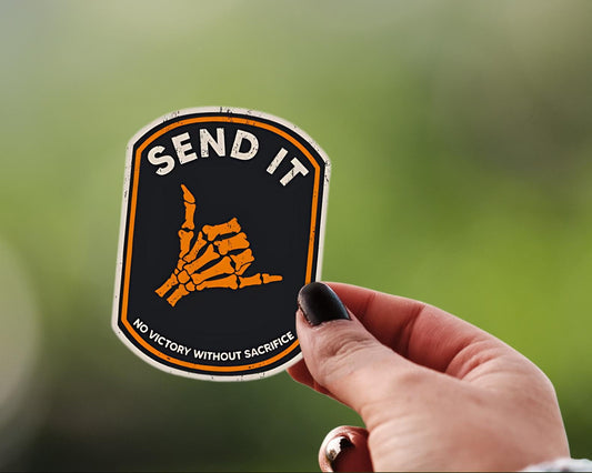 A hand with dark nail polish holds a shield-shaped sticker that reads 'SEND IT' with an orange skeletal hand graphic and the phrase 'NO VICTORY WITHOUT SACRIFICE' against a blurred green outdoor background.